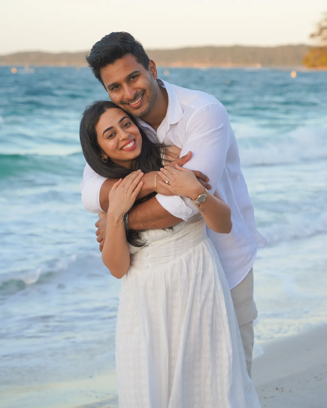 Smiling couple hugging on the beach in white outfits during an engagement shoot