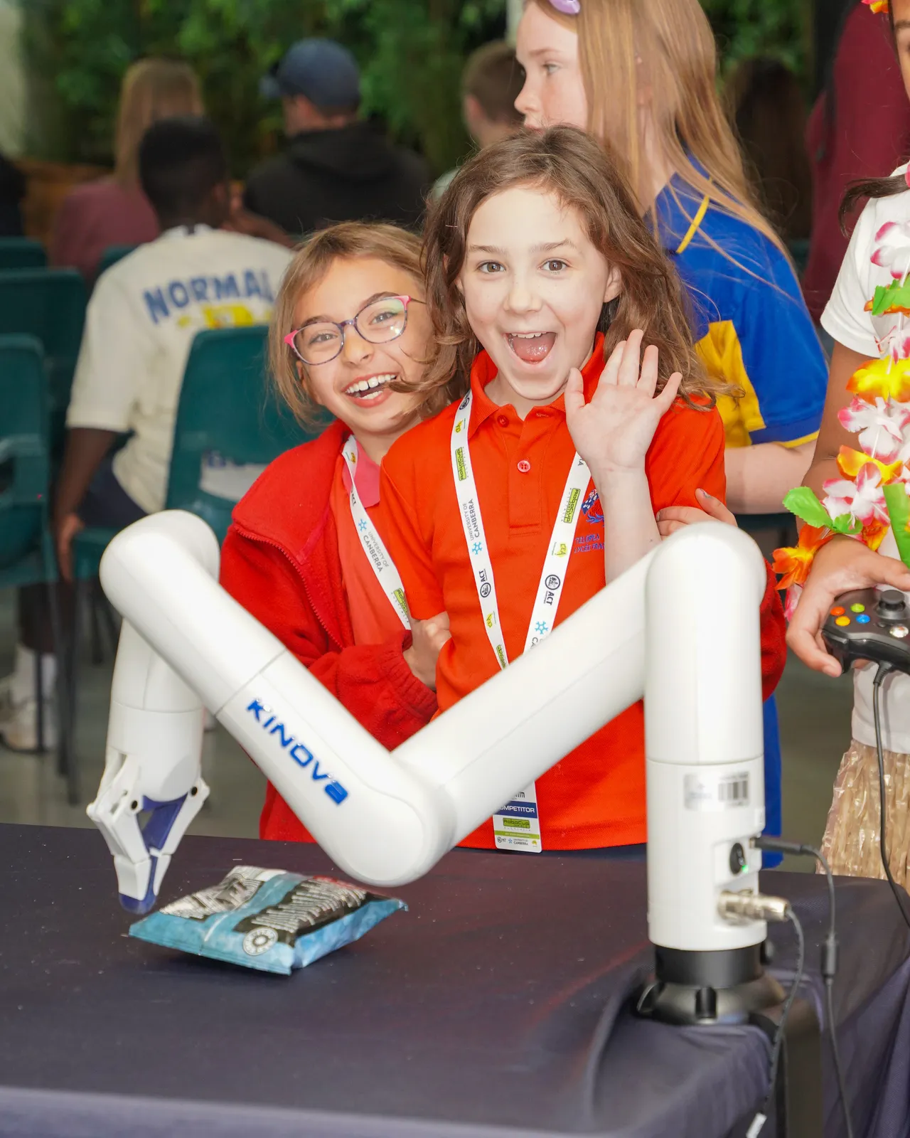 Two excited children in bright uniforms beside a robotic arm at a Canberra STEM event